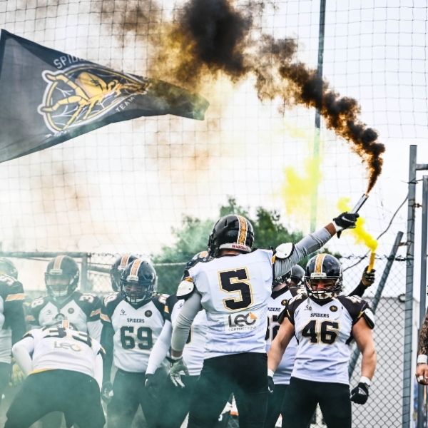 Squadra di football americano Spiders che esulta in campo con fumogeni colorati e bandiera del club.